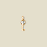 Gold key-shaped pendant with a heart shaped clear gemstone on a beige background