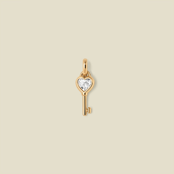 Gold key-shaped pendant with a heart shaped clear gemstone on a beige background