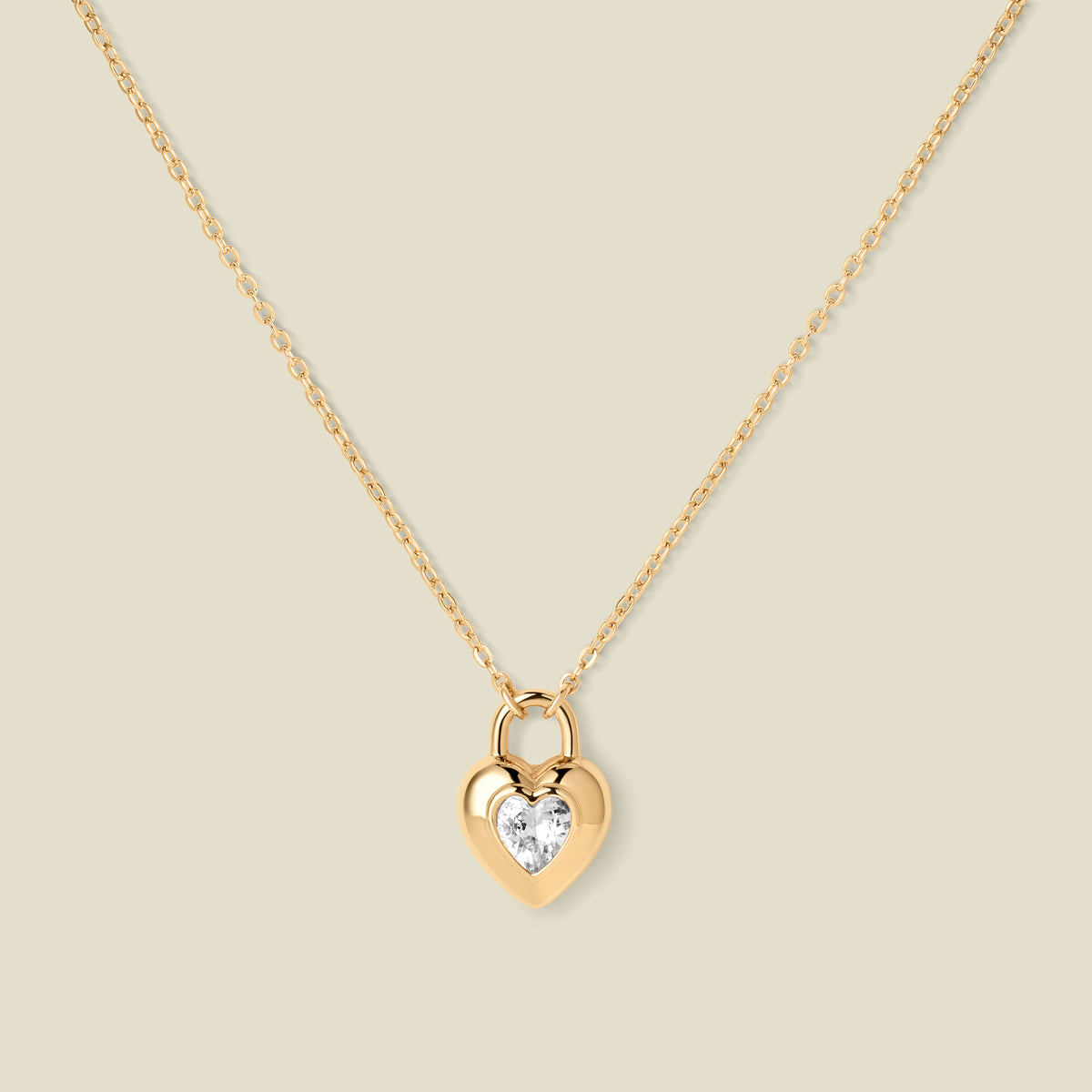 A gold heart-shaped pendant necklace with a clear heart-shaped gemstone in the center, suspended from a delicate gold chain against a solid beige background.