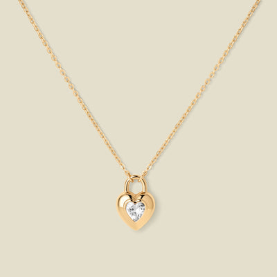 A gold heart-shaped pendant necklace with a clear heart-shaped gemstone in the center, suspended from a delicate gold chain against a solid beige background.