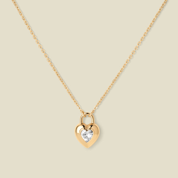 A gold heart-shaped pendant necklace with a clear heart-shaped gemstone in the center, suspended from a delicate gold chain against a solid beige background.