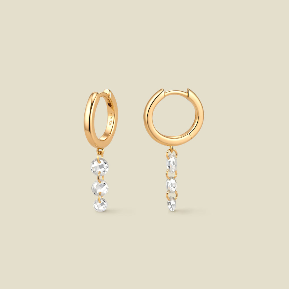 A pair of gold hoop earrings with a column of three clear round crystals dangling vertically from each, shown on a solid beige background.
