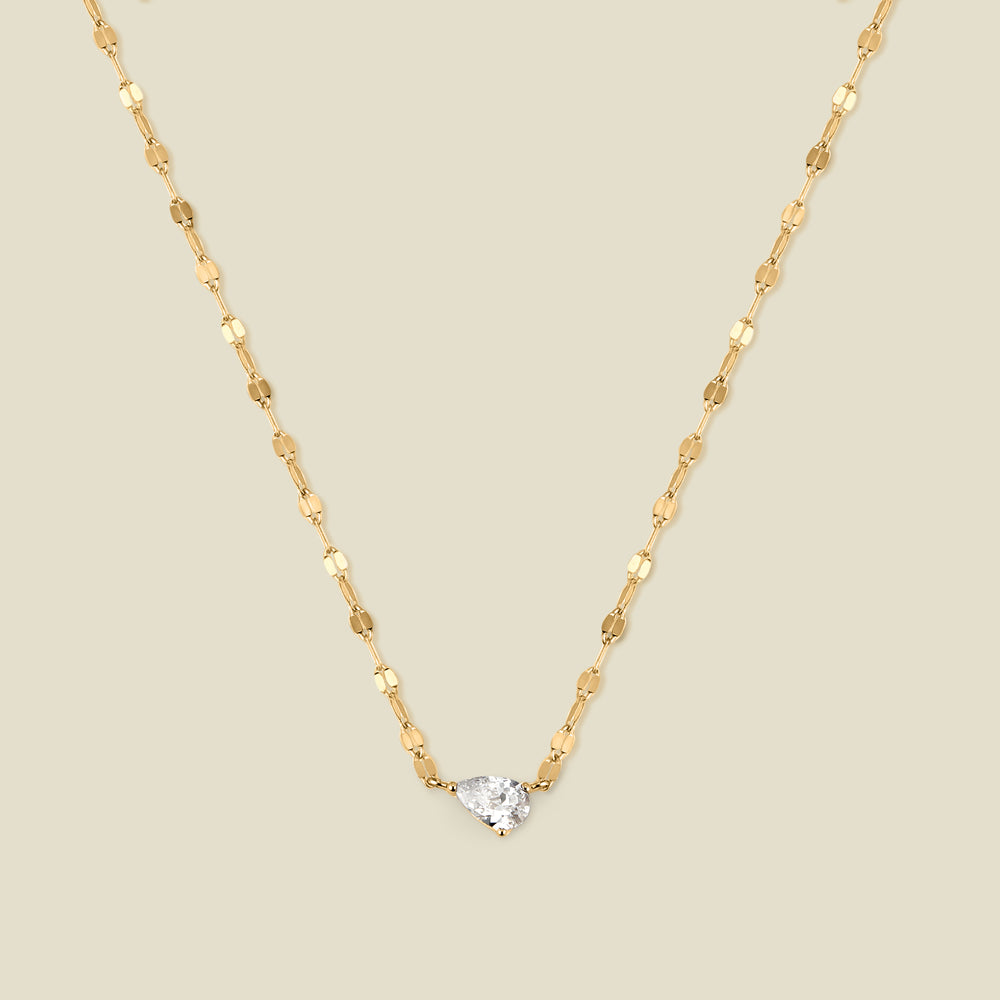 A dainty textured gold chain necklace with a solitaire tear-drop shaped clear stone, set sideways in the center of the chain, shown against a solid beige background.