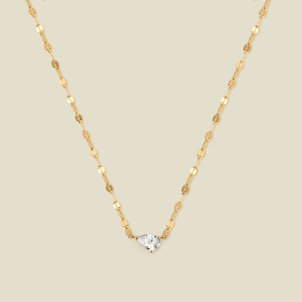 A dainty textured gold chain necklace with a solitaire tear-drop shaped clear stone, set sideways in the center of the chain, shown against a solid beige background.