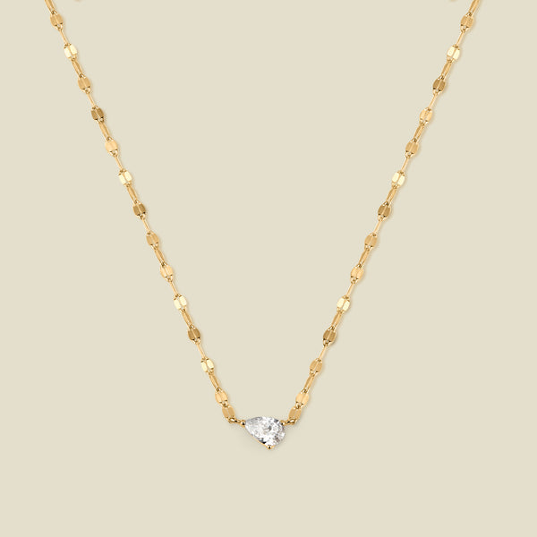 A dainty textured gold chain necklace with a solitaire tear-drop shaped clear stone, set sideways in the center of the chain, shown against a solid beige background.