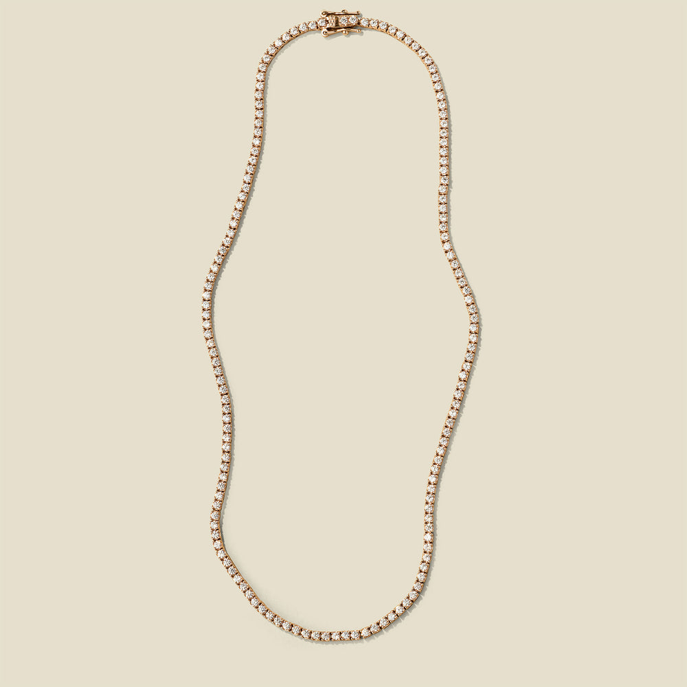 A delicate gold tennis-style chain necklace with a matching clasp, displayed on a solid light beige background.