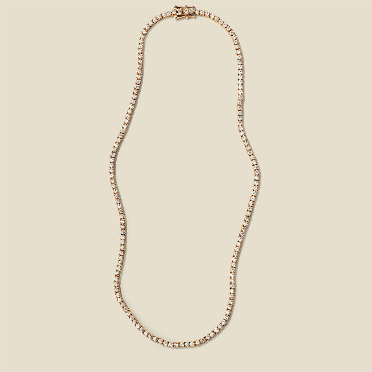 A delicate gold tennis-style chain necklace with a matching clasp, displayed on a solid light beige background.