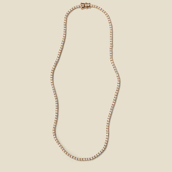 A delicate gold tennis-style chain necklace with a matching clasp, displayed on a solid light beige background.