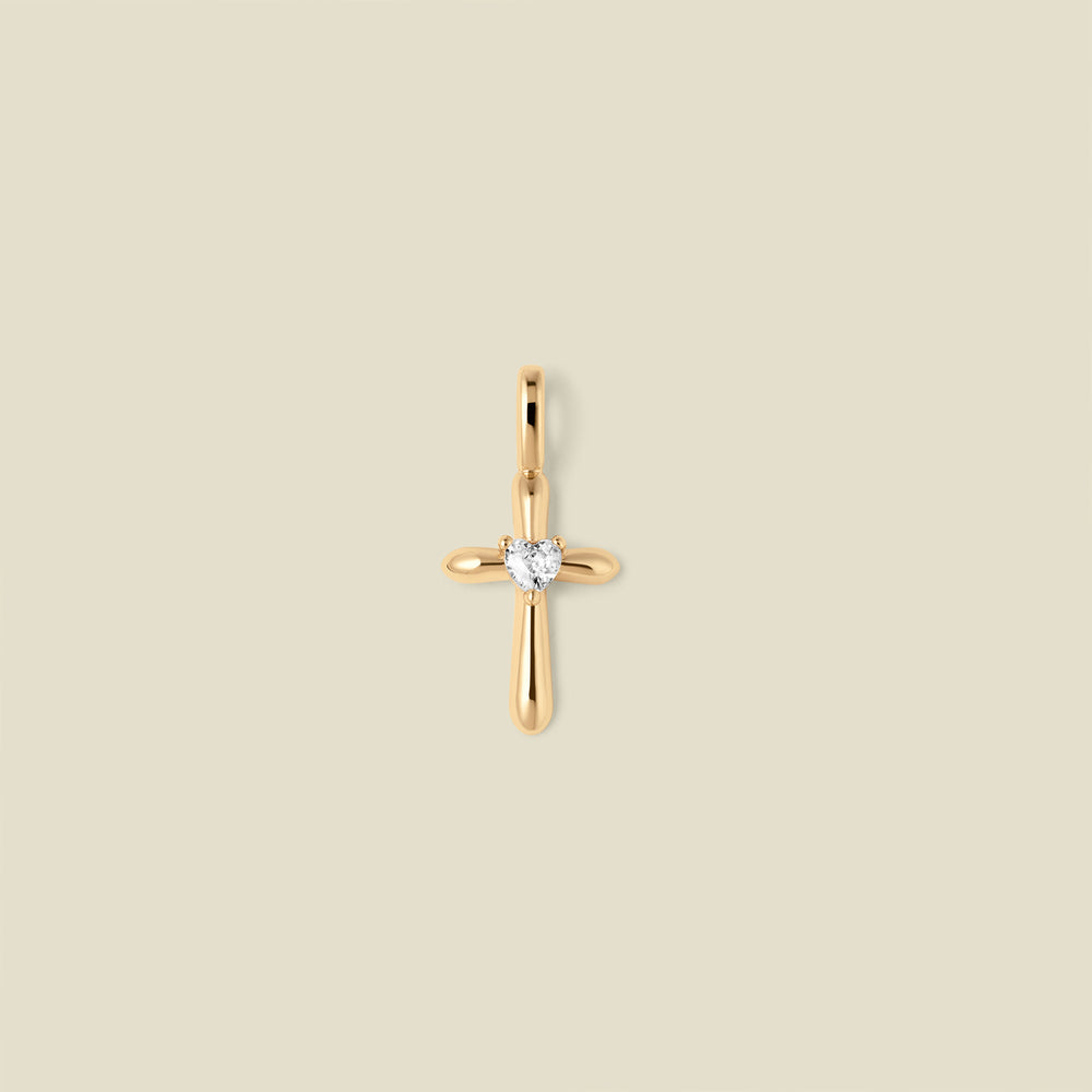 A polished gold cross pendant with a single clear heart-shaped gemstone at the center, shown against a solid light beige background.