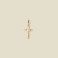 A polished gold cross pendant with a single clear heart-shaped gemstone at the center, shown against a solid light beige background.