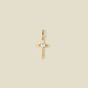 A polished gold cross pendant with a single clear heart-shaped gemstone at the center, shown against a solid light beige background.
