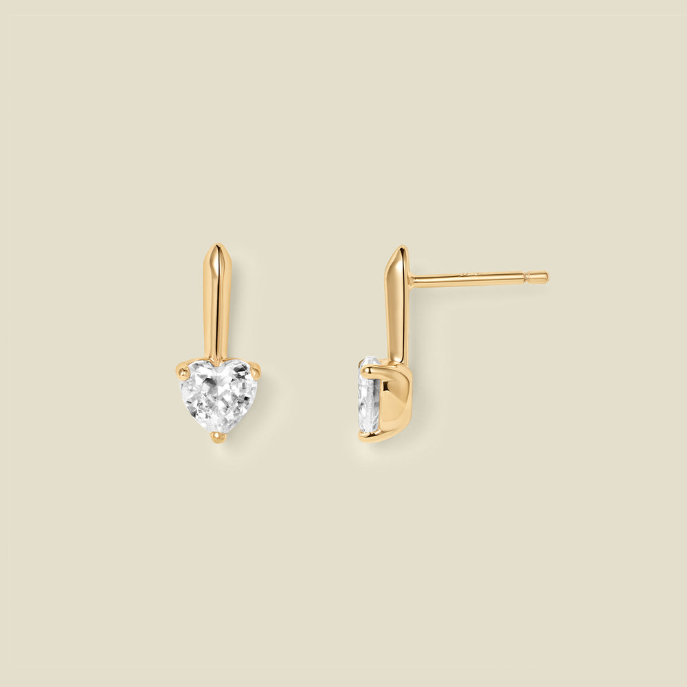 A pair of gold drop earrings with heart-shaped clear gemstones, shown from front and side angles on a solid beige background.