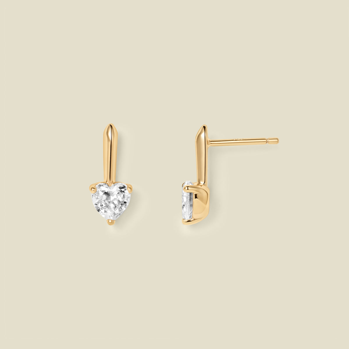 A pair of gold drop earrings with heart-shaped clear gemstones, shown from front and side angles on a solid beige background.