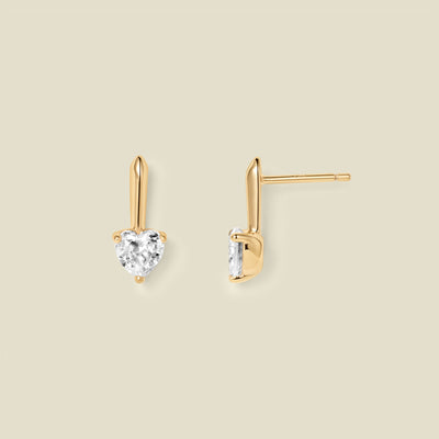 A pair of gold drop earrings with heart-shaped clear gemstones, shown from front and side angles on a solid beige background.