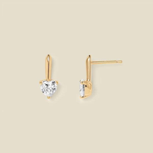 A pair of gold drop earrings with heart-shaped clear gemstones, shown from front and side angles on a solid beige background.