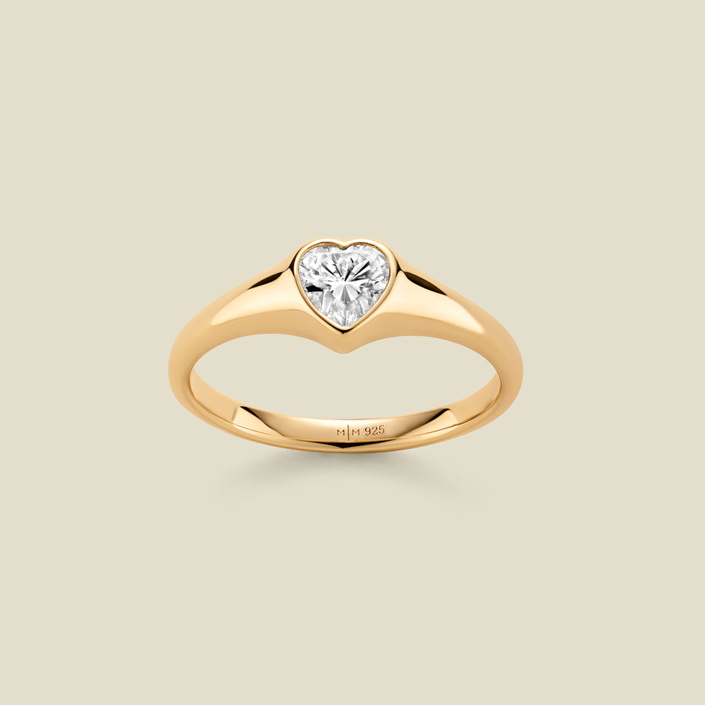A heart-shaped clear gemstone signet ring in a yellow gold setting, centered on a light beige background. 