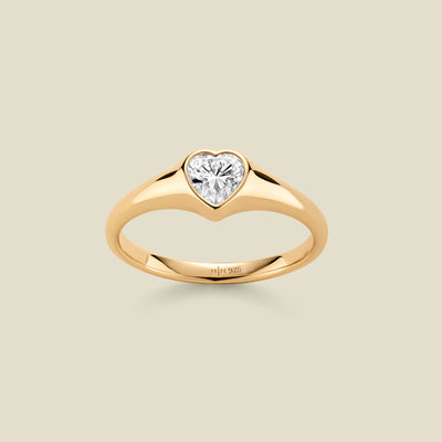 A heart-shaped clear gemstone signet ring in a yellow gold setting, centered on a light beige background. 
