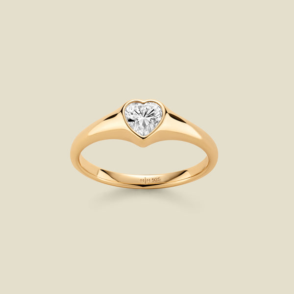 A heart-shaped clear gemstone signet ring in a yellow gold setting, centered on a light beige background. 