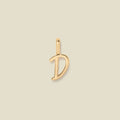 A polished gold initial pendant charm shaped like the letter 'D' in a cursive script, centered on a solid light beige background.