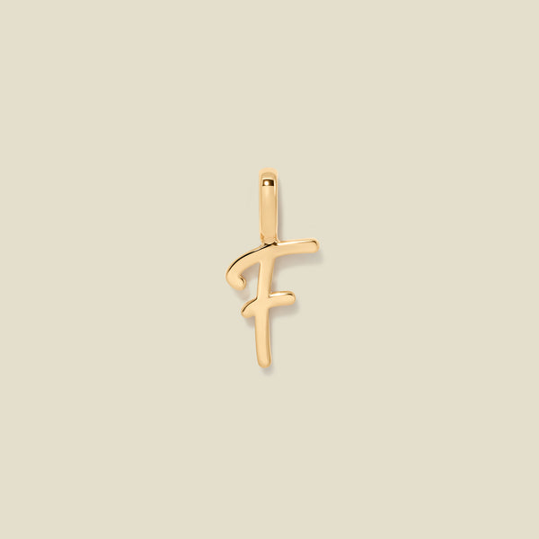 A polished gold initial pendant charm shaped like the letter 'F' in a cursive script, centered on a solid light beige background.