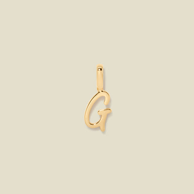 A polished gold initial pendant charm shaped like the letter 'G' in a cursive script, centered on a solid light beige background.