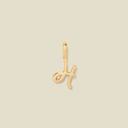 A polished gold initial pendant charm shaped like the letter 'H' in a cursive script, centered on a solid light beige background.