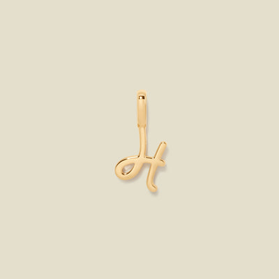 A polished gold initial pendant charm shaped like the letter 'H' in a cursive script, centered on a solid light beige background.