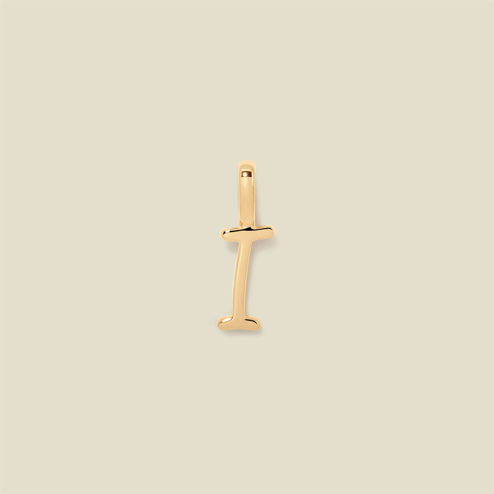 A polished gold initial pendant charm shaped like the letter 'I' in a cursive script, centered on a solid light beige background.