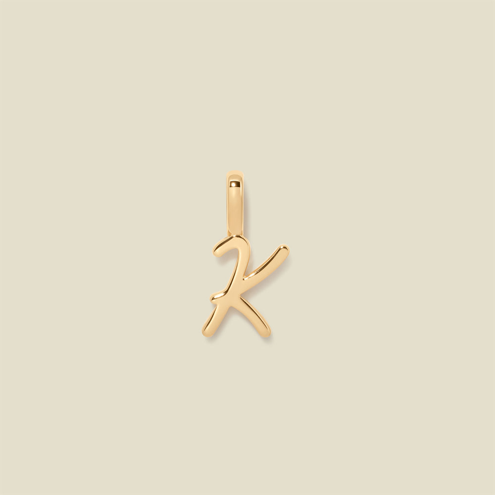 A polished gold initial pendant charm shaped like the letter 'K' in a cursive script, centered on a solid light beige background.