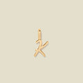 A polished gold initial pendant charm shaped like the letter 'K' in a cursive script, centered on a solid light beige background.