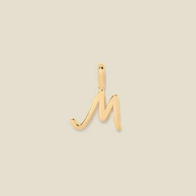 A polished gold initial pendant charm shaped like the letter 'M' in a cursive script, centered on a solid light beige background.