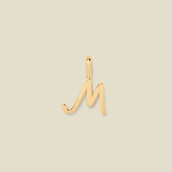 A polished gold initial pendant charm shaped like the letter 'M' in a cursive script, centered on a solid light beige background.