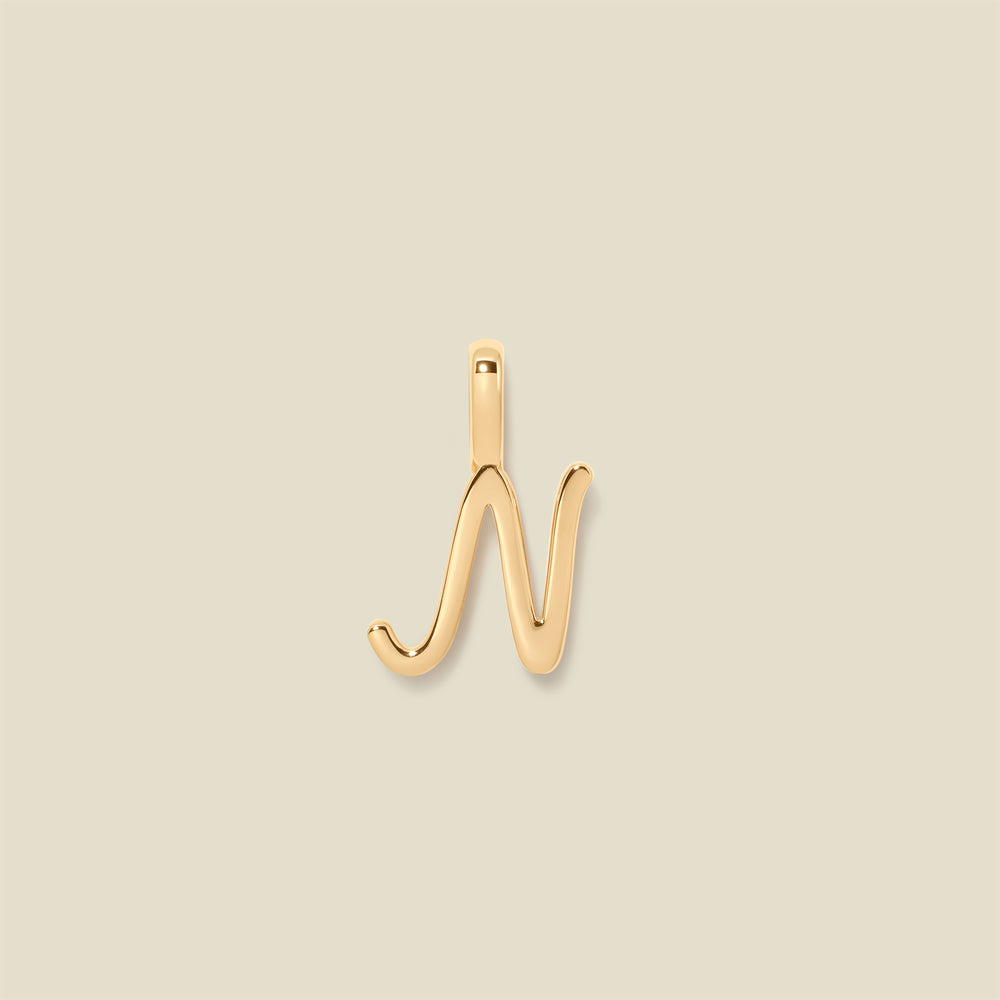 A polished gold initial pendant charm shaped like the letter 'N' in a cursive script, centered on a solid light beige background.