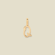 A polished gold initial pendant charm shaped like the letter 'Q' in a cursive script, centered on a solid light beige background.