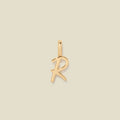 A polished gold initial pendant charm shaped like the letter 'R' in a cursive script, centered on a solid light beige background.
