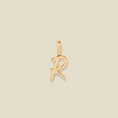 A polished gold initial pendant charm shaped like the letter 'R' in a cursive script, centered on a solid light beige background.