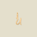 A polished gold initial pendant charm shaped like the letter 'U' in a cursive script, centered on a solid light beige background.