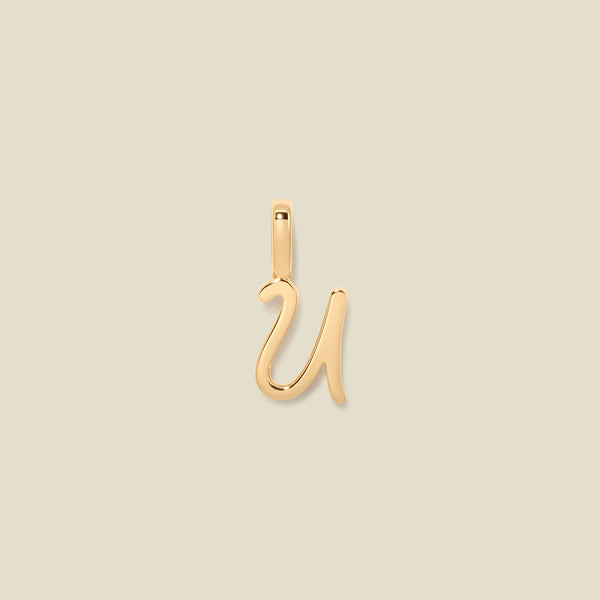 A polished gold initial pendant charm shaped like the letter 'U' in a cursive script, centered on a solid light beige background.