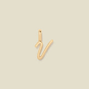 A polished gold initial pendant charm shaped like the letter 'V' in a cursive script, centered on a solid light beige background.