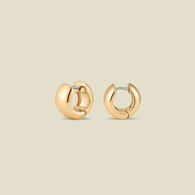 A pair of wide, polished gold huggie hoop earrings standing up right at two different angles, set against a solid beige background.