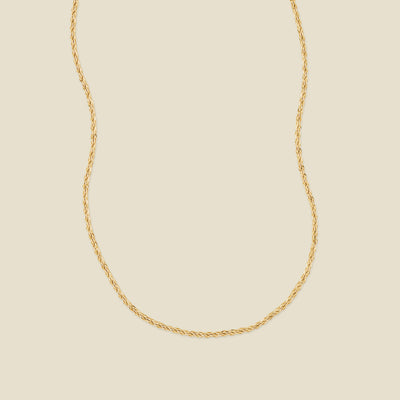 A delicate gold chain necklace with a twisted rope texture, displayed against a solid light beige background.