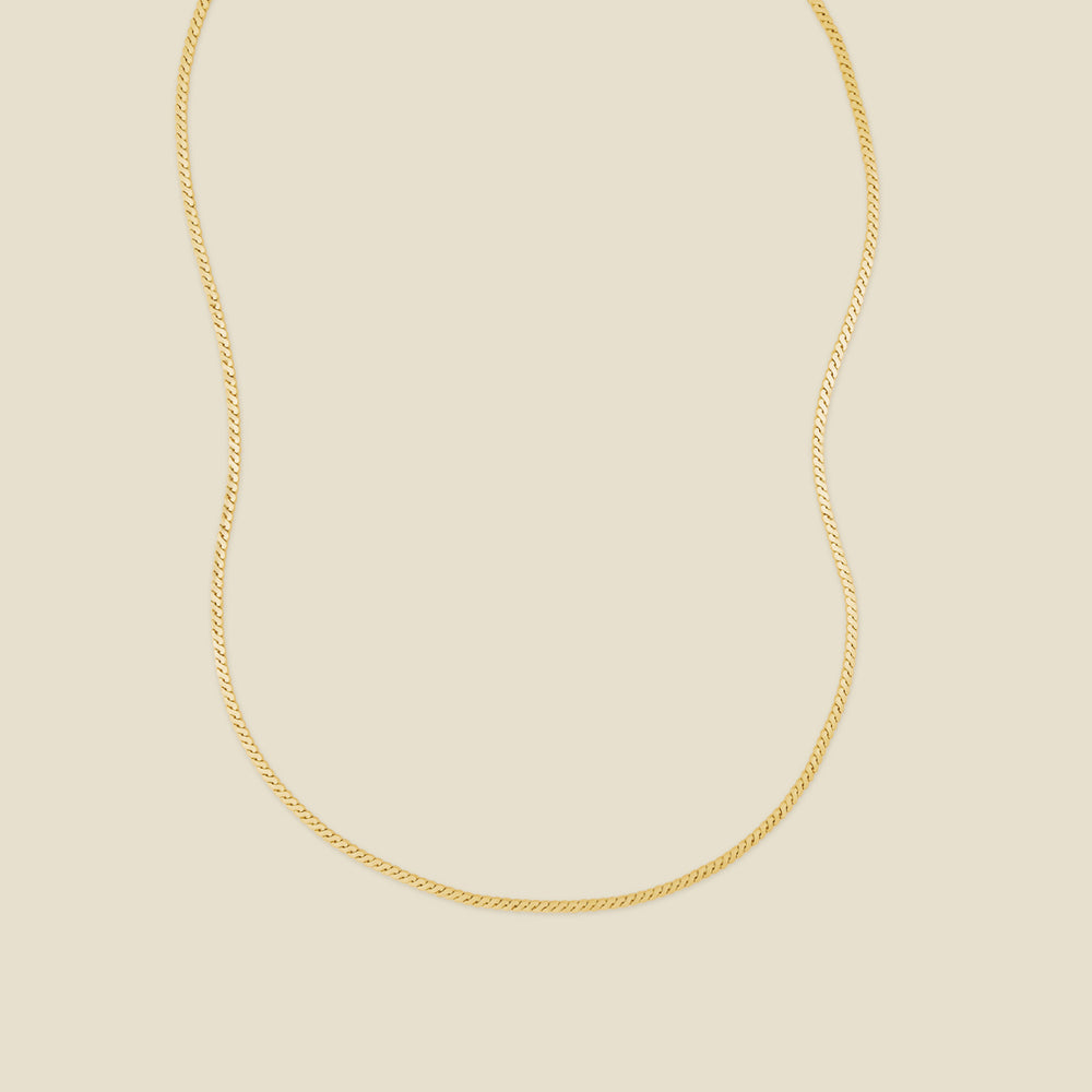 A gold flat, braided chain necklace set against a solid beige background.