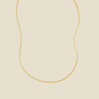A gold flat, braided chain necklace set against a solid beige background.