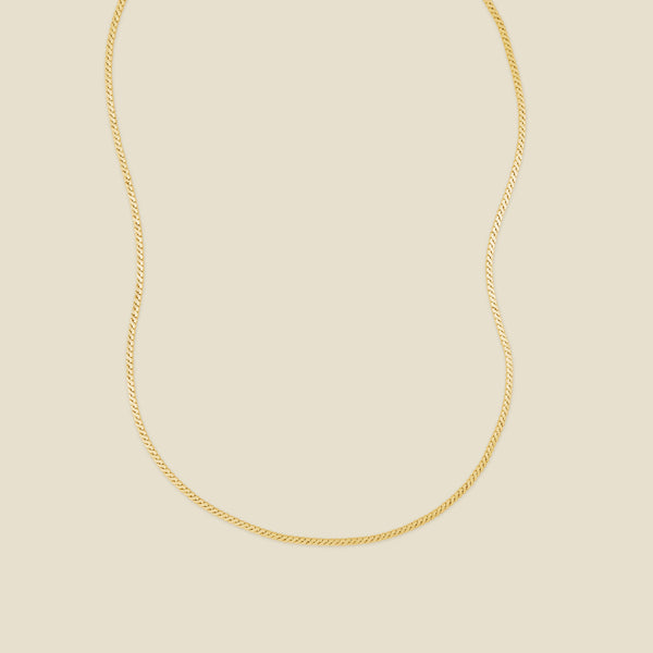 A gold flat, braided chain necklace set against a solid beige background.