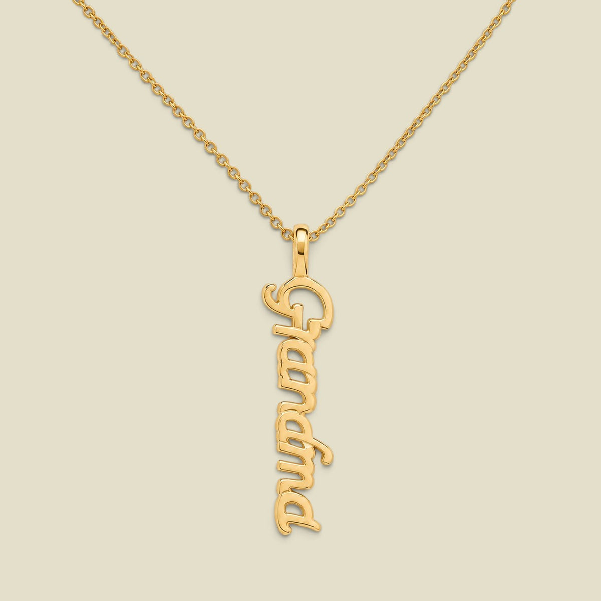 A gold script nameplate necklace spelling 'Grandma'  hanging vertically on a delicate gold chain, presented on a solid light beige background.