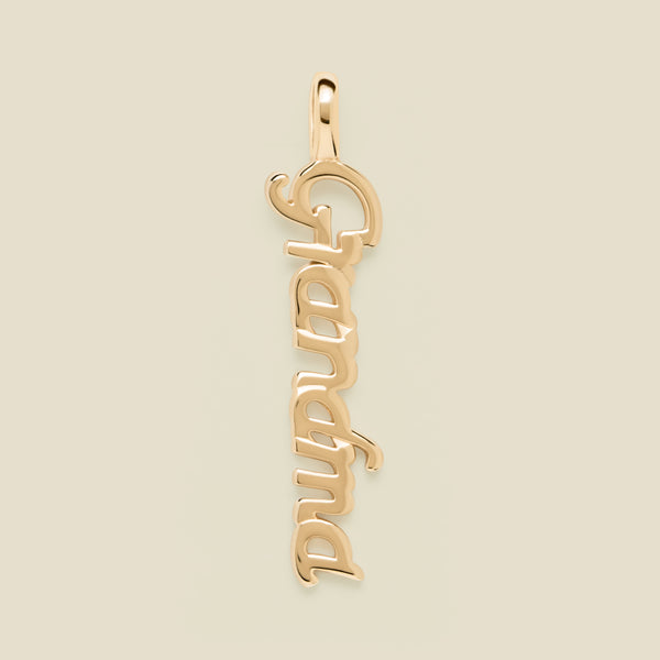 A polished gold script pendant spelling 'Grandma' vertically, shown on a solid light beige background.