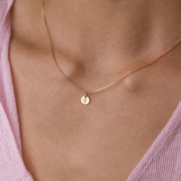 A delicate gold chain necklace with a small, flat circular disc pendant featuring a tiny engraved cactus, worn on a model's neck over a light pink knit top.