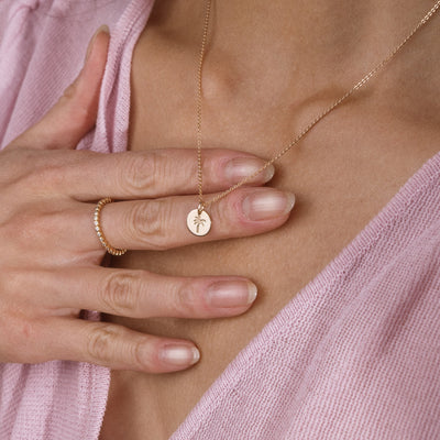 A close-up of a person wearing a delicate gold necklace with a small round pendant engraved with a palm tree and a gold ring, styled with a pink knit top.