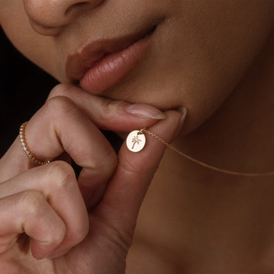 A close-up lifestyle shot of a model holding a delicate gold chain necklace with a small round disc pendant engraved with a palm tree, while wearing a gold ring.