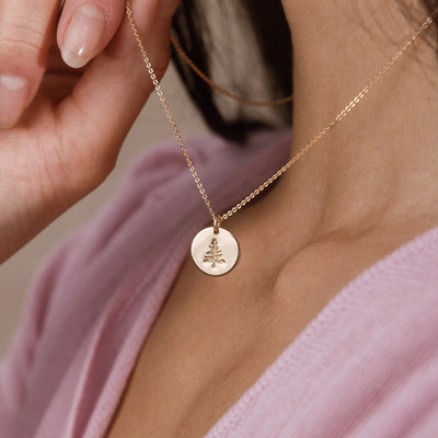 A close-up lifestyle shot of a model wearing a delicate gold chain necklace with a small round disc pendant engraved with a pine tree, styled with a soft pink top.